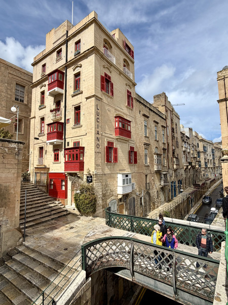 Jackson Rodriguez — A charming street scene in Valletta with traditional limestone buildings, vivid red wooden balconies, and pedestrians crossing a small bridge above a narrow road.