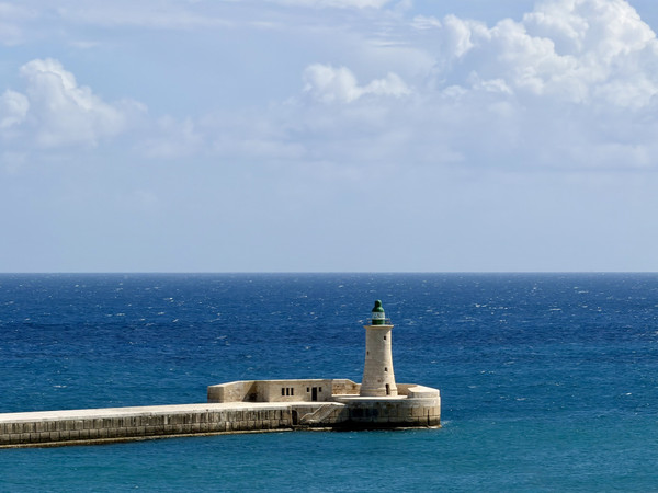 Jackson Rodriguez — A limestone lighthouse and keeper’s cottage perch at the end of a long stone breakwater jutting into a vivid cobalt-blue Mediterranean sea. Velleta- Malta 🇲🇹