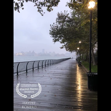 Lamplight glistens on the wet planks of the Boardwalk at Jersey City one recent rainy morning. The buildings of Manhattan loom out of low-lying clouds in the background.