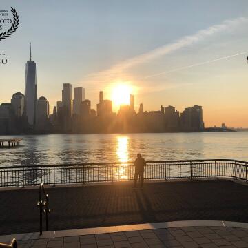 A person watches as the sun rises over lower Manhattan, sending a blade of gold across the Hudon river toward Jersey City. In this photo, the lines of the sunshine, of the shadows of New York's skyscrapers on the water, of the railings and of the light shining through the railings draw in the eye. <br />
