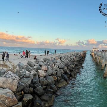 People walk out on the pier and adjacent tidal barrier at South Pointe Park in Miami Beach as the setting sun casts evening colors on a line of clouds.