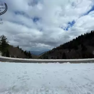 This picture was taken on a cold February day high up in the Great Smoky Mountains National Park, where a graceful curve in the road frames the puffy clouds in the bright blue sky and the mountains beyond, and the bumps and tufts of piled up snow on the ground.