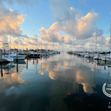Clouds are reflected in the tranquil water between watercraft as the sun rises and puffy clouds gather over Coconut Grove marina in Miami, Florida.