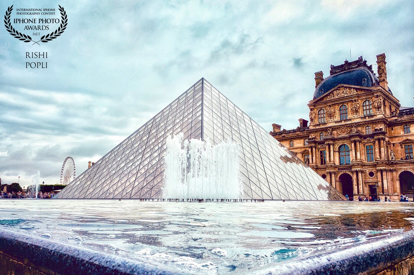 During my recent travel to Paris, I found myself walking the streets on a cloudy day trying to capture some great shots. This photograph was taken in the center of the Louvre Museum, with the backdrop of the famous Pyramid and architectural historical buildings.  