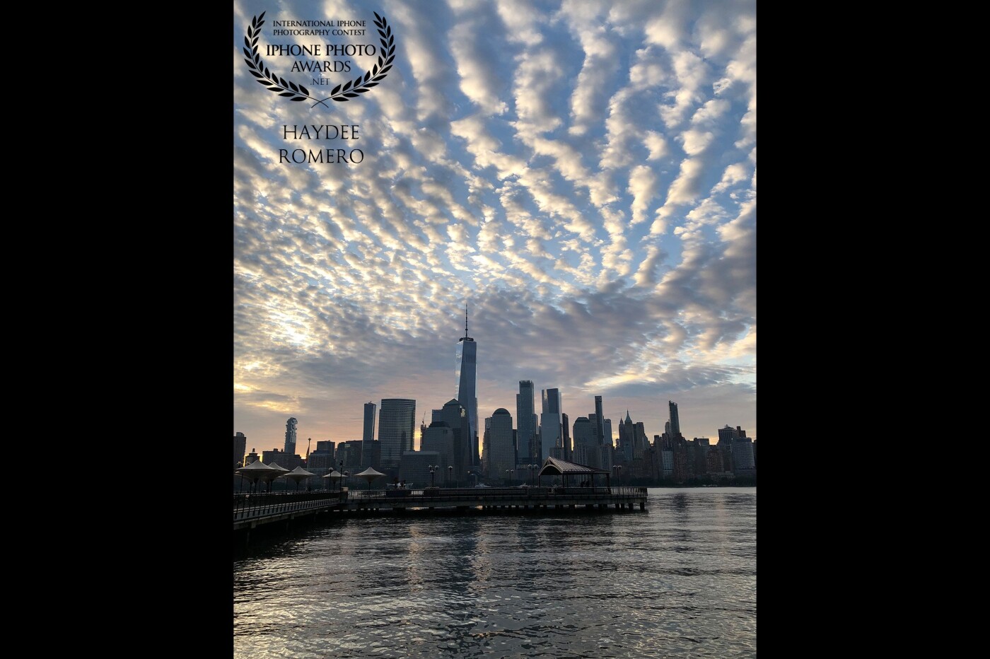 In this photo, the morning sun reflects off wavy and puffy lines of clouds over the buildings of lower Manhattan and onto the waters of the Hudson River off Jersey City. 