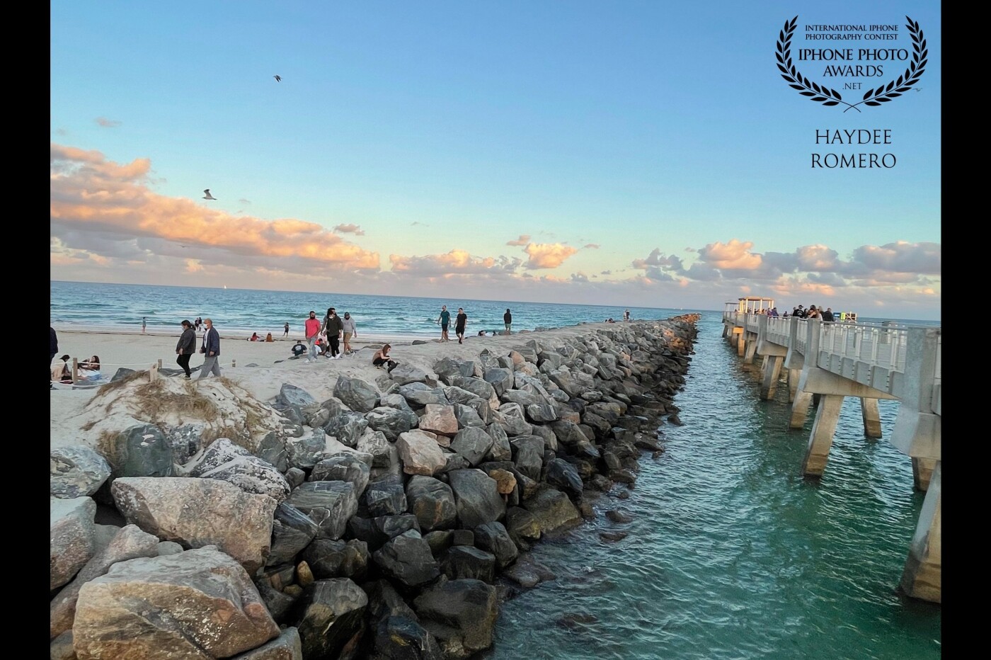 People walk out on the pier and adjacent tidal barrier at South Pointe Park in Miami Beach as the setting sun casts evening colors on a line of clouds.