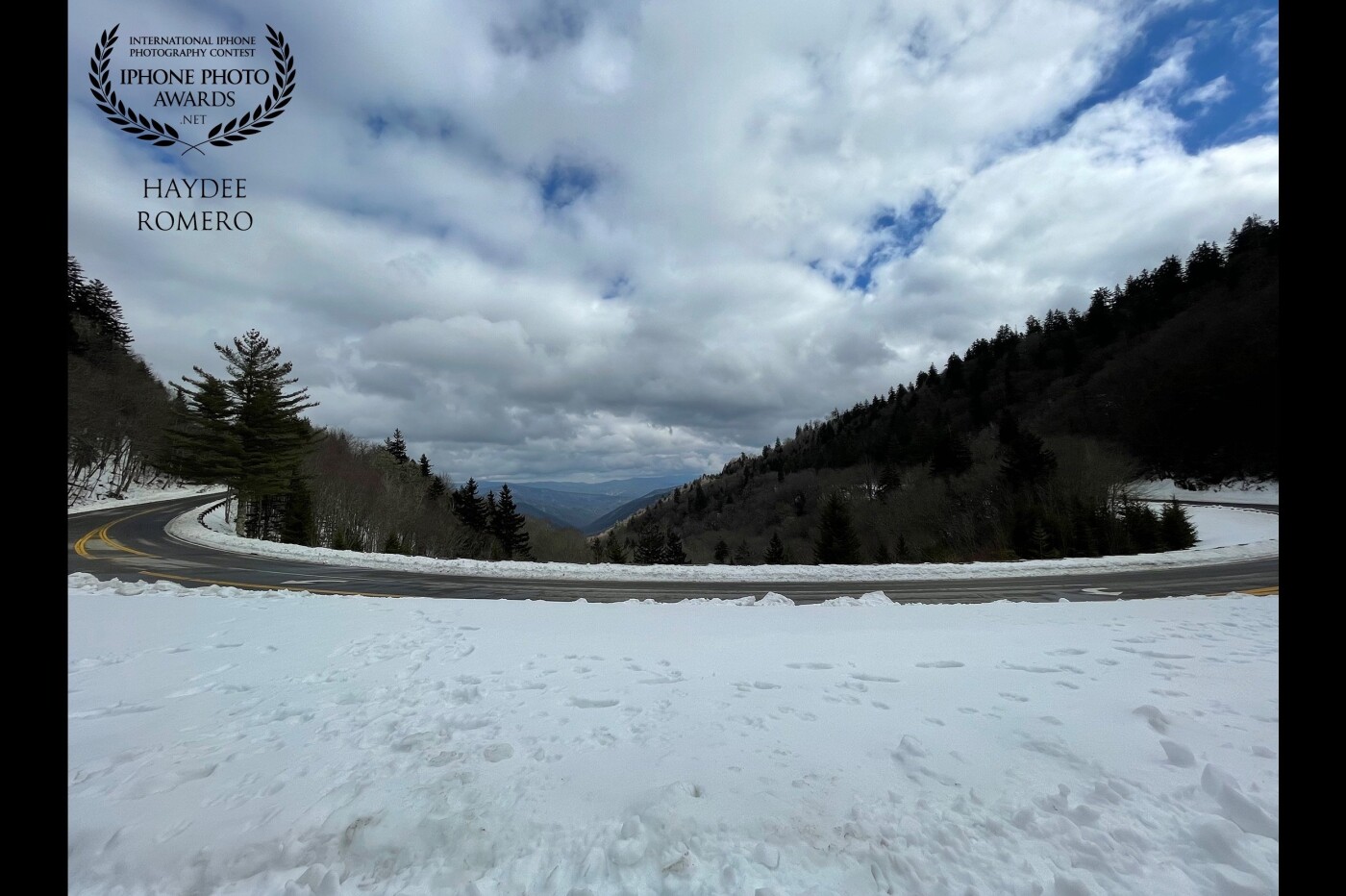This picture was taken on a cold February day high up in the Great Smoky Mountains National Park, where a graceful curve in the road frames the puffy clouds in the bright blue sky and the mountains beyond, and the bumps and tufts of piled up snow on the ground.