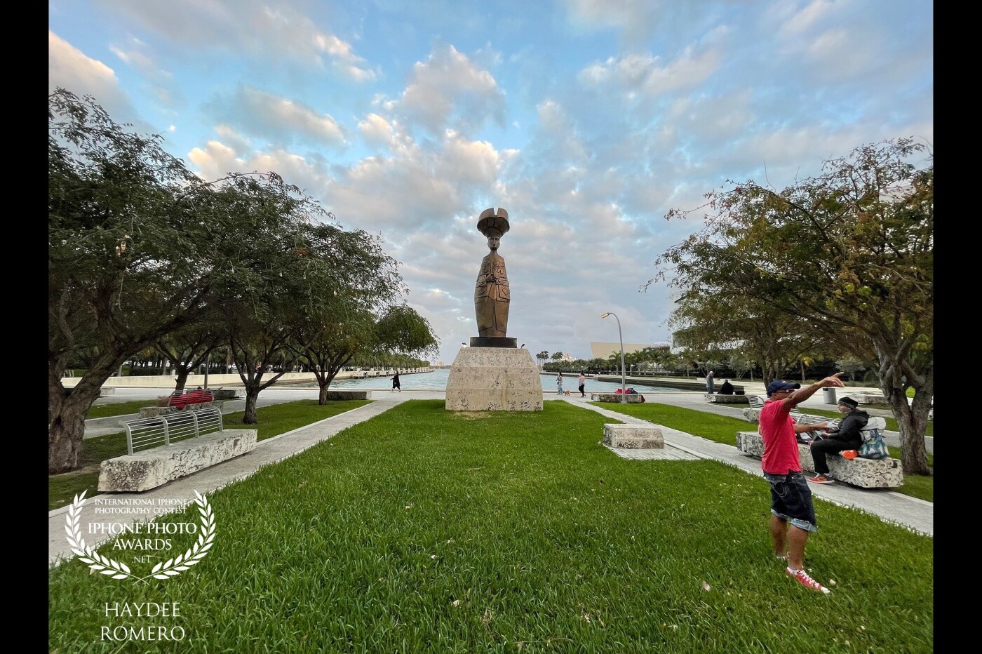 Clouds frame a statue dedicated to Flora, the Collector of Dreams, by the renowned Cuban artist Cundo Bermudez, on the waterfront Maurice A. Ferre Park in downtown Miami, where the stone benches are occupied by the homeless while city residents stroll, run, cycle or  walk their dogs along the bayfront.