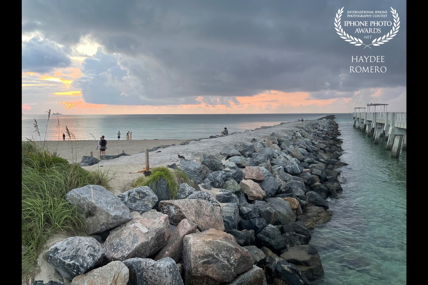 The opening sun ponds though tropical storm clouds off Miami Beach while tourists walk on the sand, take photos of daybreak or sit on the stones of a rock jetty at South Pointe Park in Miami Beach.