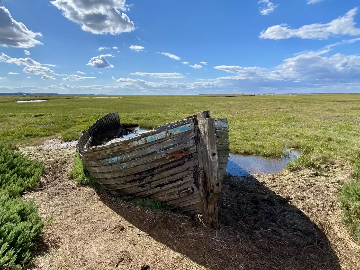 The old wooden boat resting in a grassy marsh under  a bright blue sky with scattered clouds. Blaken...