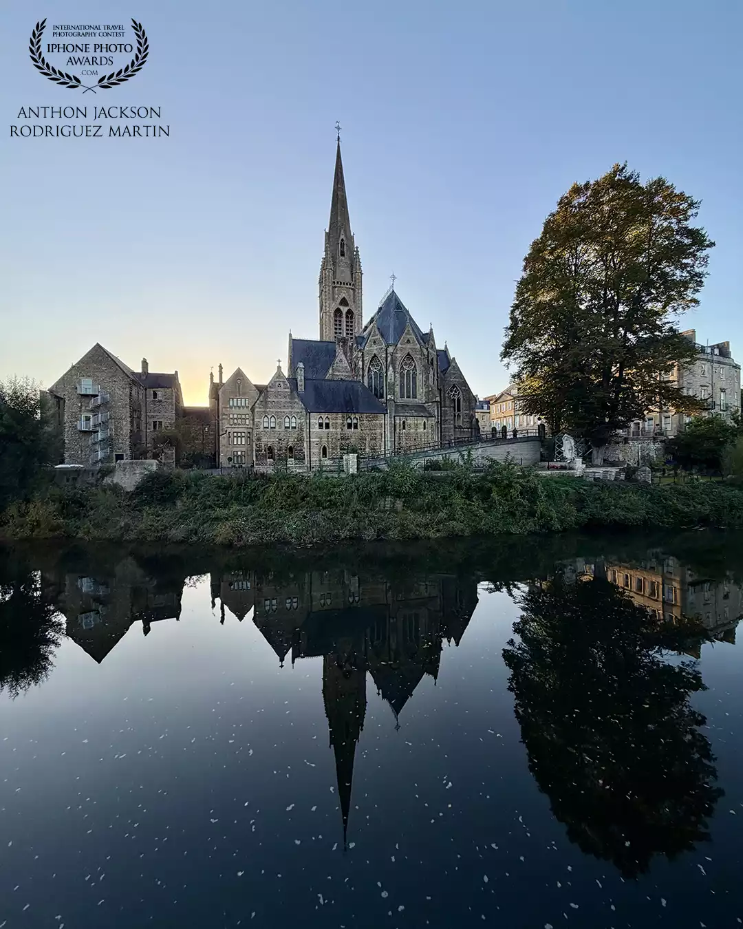 The beautiful St John's Church in Bath with the tall steeple reflects in the calm river at the golden hour, surrounded by trees and amazing old architecture.