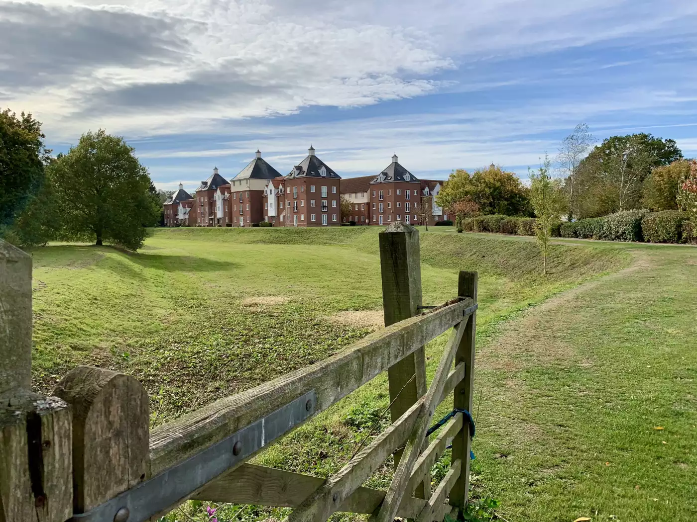 A peaceful green landscape with a wooden gate in the foreground, leading toward a complex of red-bri...