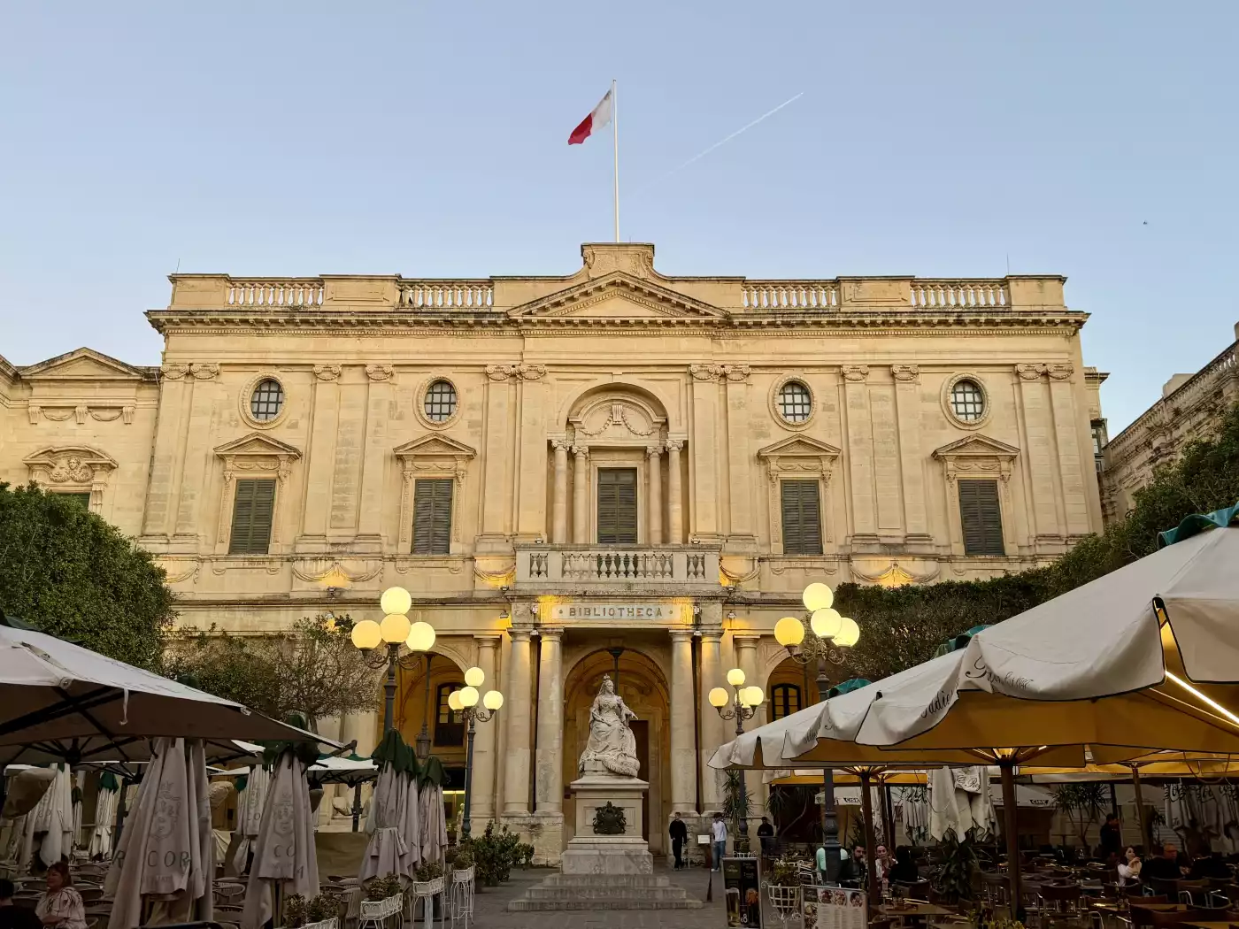 Malta’s National Library in Valletta — glows warmly at dusk, with a statue of Queen Victoria in fron...