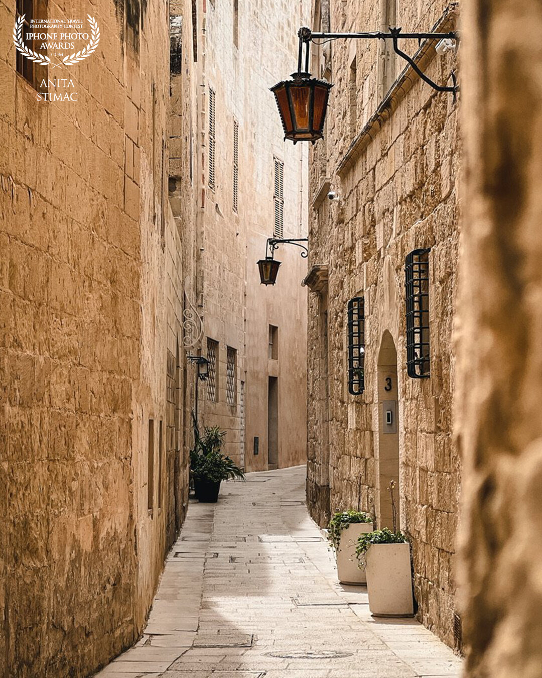 A quiet, narrow street in the historic town of Mdina, Malta. The rough, stone pavement and empty alleyway evoke a sense of solitude and history. Above, a lone lantern hangs silently, a reminder of the street's past.