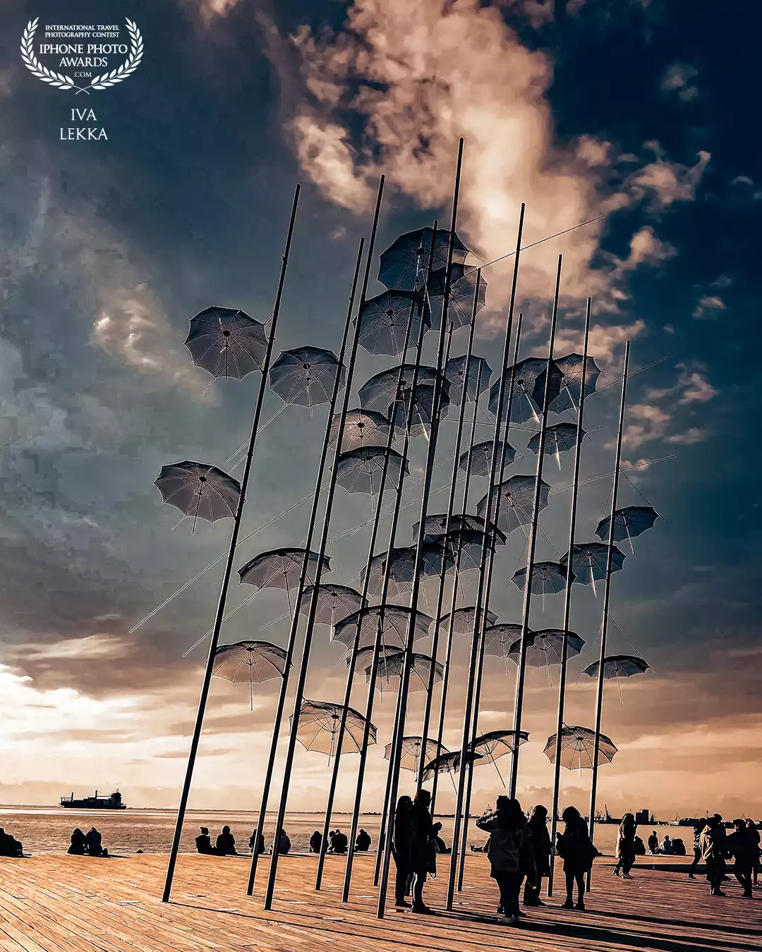 Umbrellas Installation, Thessaloniki Seafront.<br />
The Umbrellas installation stands quietly by the sea in Thessaloniki, tall, delicate, and a little surreal. Created by Greek sculptor Giorgos Zongolopoulos, the piece is open to interpretation: some see protection, others freedom, or a sense of lightness. For me, it reflects the mood of the city itself: creative, open, and always aware of its past and present. I captured this moment as the sun was setting, when people began to slow down and gather underneath. It felt like a shared pause, a space between movement and stillness that Thessaloniki naturally holds so well.